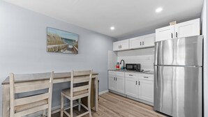 Une cuisine moderne avec des meubles blancs, un réfrigérateur en acier inoxydable, une table à manger en bois avec des chaises, et une décoration murale sur le thÚme de la plage.