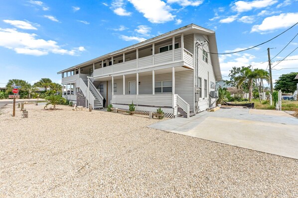 A two-story building with a balcony and a covered porch, located on a gravel lot with palm trees and a stop sign in the background. - 🐾 Dog Friendly, Spacious Beach Retreat, only Steps to Sugary Sand, Conveniences and all the Fun! (Fort Myers Beach)