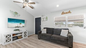 Un salon avec un canapé gris foncé, une console blanche, un ventilateur de plafond, un tableau représentant une scÚne de plage et un panneau mural sur lequel on peut lire le mot « Plage ».