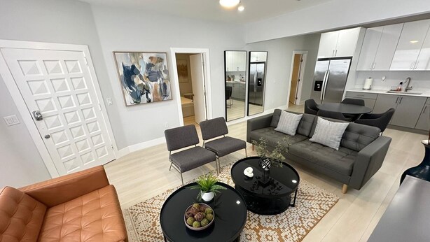 A modern living room with a leather sofa, two black coffee tables, and a grey sofa with cushions. There are two grey chairs, a patterned rug, and a large mirror reflecting the kitchen area.