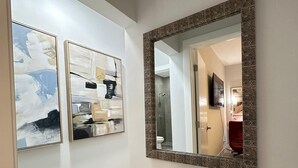 A hallway with two framed abstract paintings on the wall, a large mirror with a patterned frame, and a light-colored wooden floor.