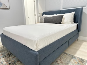 Ein Bett mit grauem gepolsterten Kopfteil, weißer Bettwäsche und einem gemusterten Teppich.