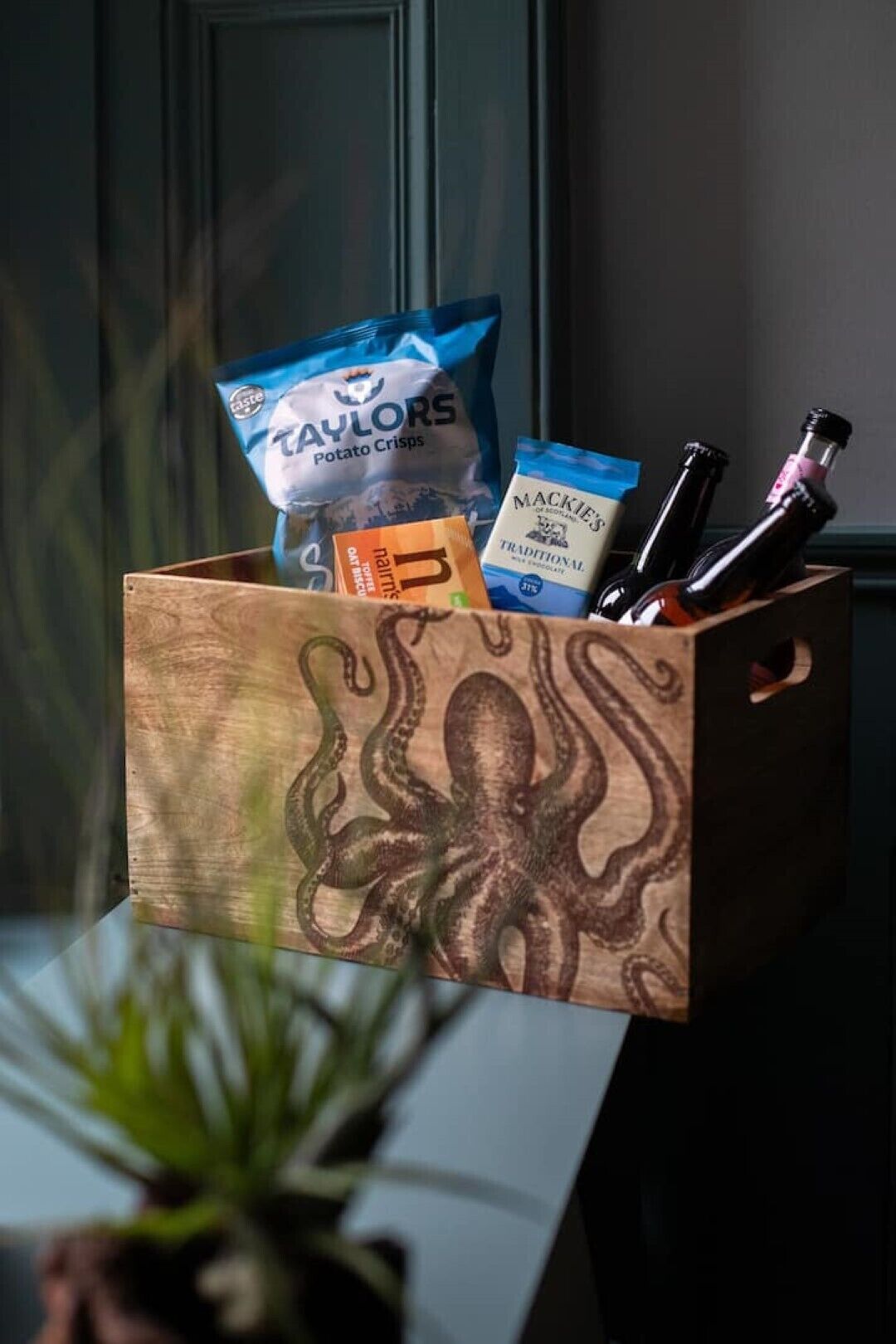 A wooden box with an octopus design, containing snacks, a bottle of wine, and a tube of cream.