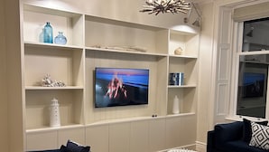 En moderne stue med et fladskærms-tv, mørke sofaer, et hvidt sofabord og indbyggede reoler med pynteting.