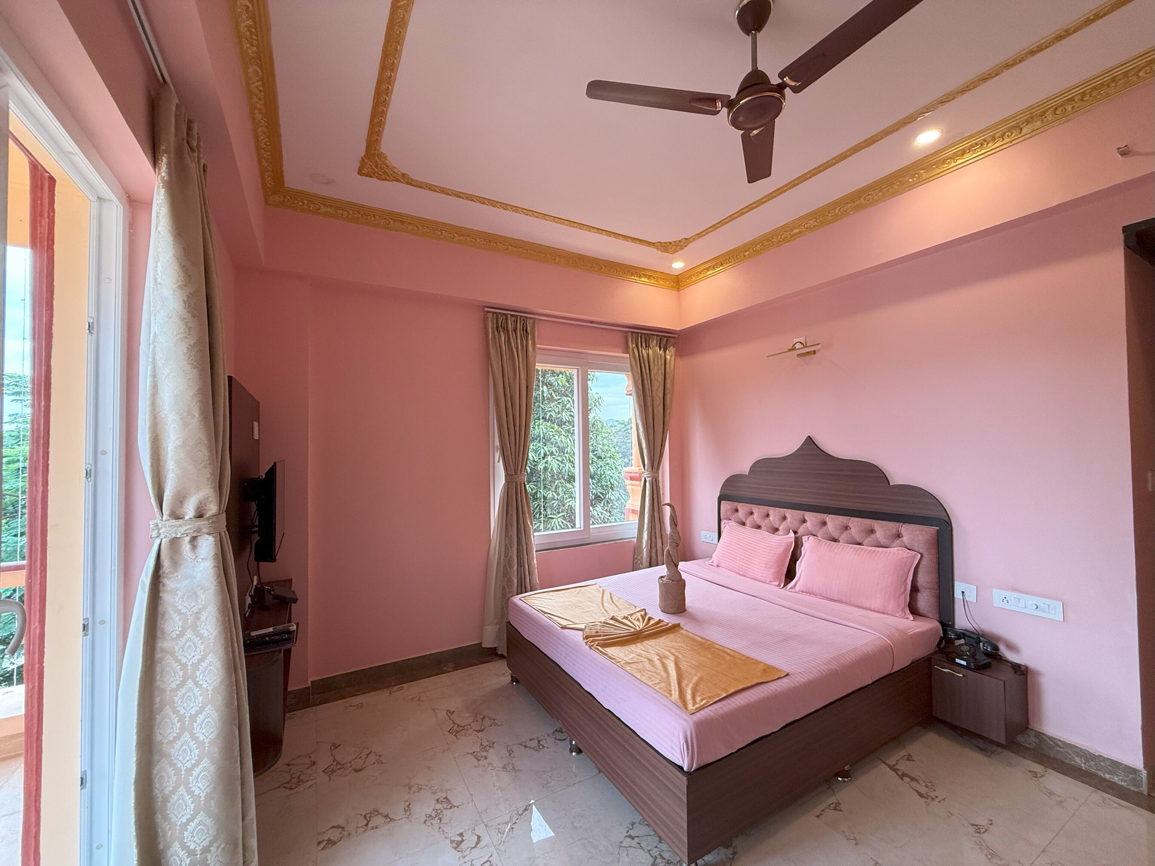 Une chambre à coucher avec un lit, un ventilateur de plafond, une fenêtre avec des rideaux et un téléviseur fixé au mur.