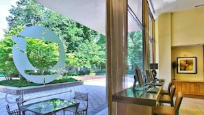 A hotel lobby with a large glass table and chairs, a computer desk with a monitor, and a view of a park with a large sculpture.