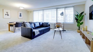 A modern living room with a blue sofa, a dining table with chairs, a large window with blinds, and a potted plant.