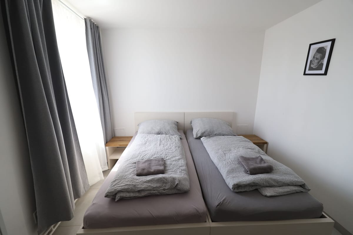A twin room with two beds, a wooden nightstand, a framed picture on the wall, and a window with curtains.