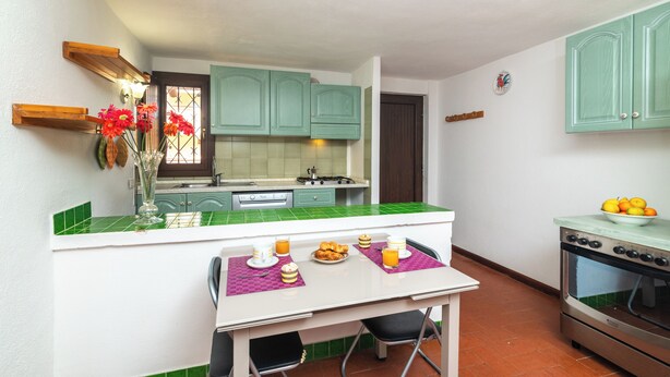 A compact kitchen with a dining area, green tiles, and a stove. - Trilo Triglia Cala Reale 229 (Golfo Aranci)