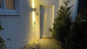 Un chemin mène à une porte, avec un petit jardin d’un côté et un buisson de l’autre.