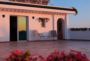 Eine Dachterrasse mit Schiebeglastür, zwei Liegestühlen, einem kleinen Tisch und Topfpflanzen.