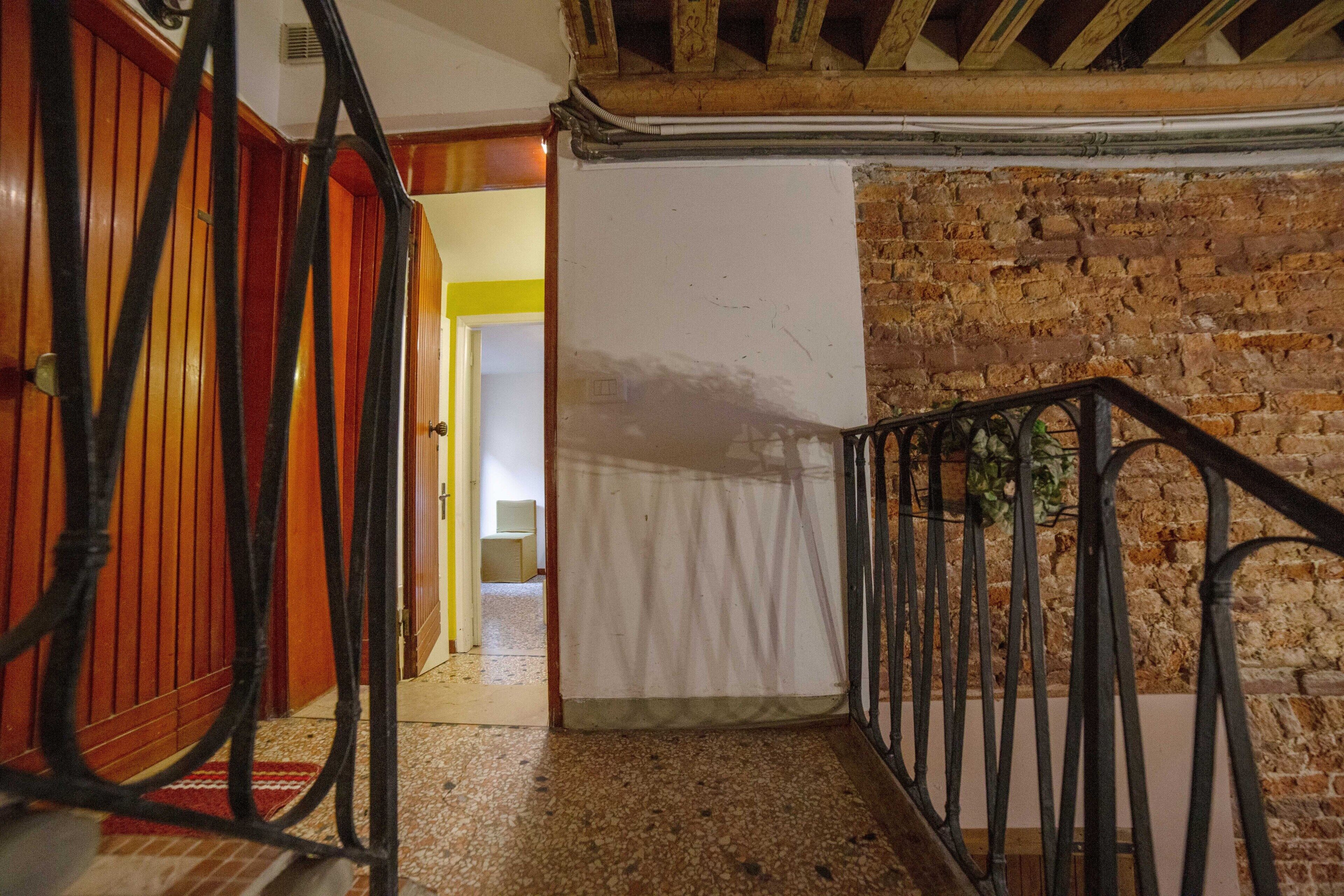 Una escala amb barana de fusta i vidre metàl·lic, que condueix a una habitació amb paret de maó i porta blanca.