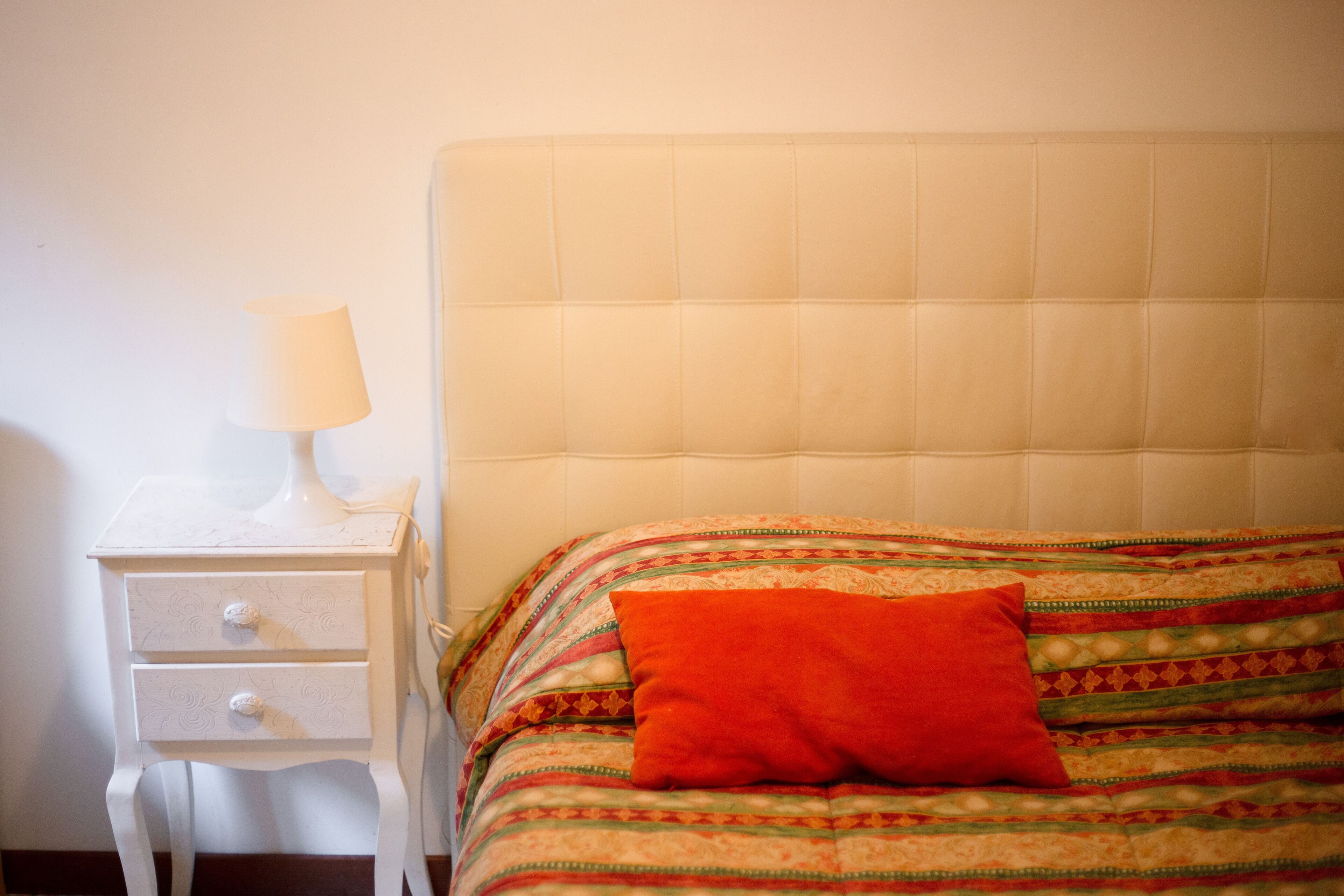 Ein Bett mit einem gemusterten Bettüberwurf und einem einzelnen orangefarbenen Kissen, ein weißer Nachttisch mit einer Lampe und einer Schublade.