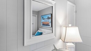 Casa de banho com toucador branco, espelho, quadro emoldurado de uma carrinha e candeeiro branco.