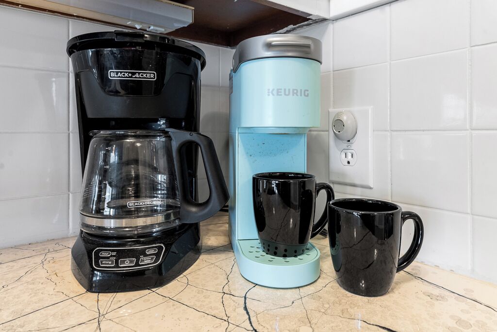 A kitchen with a black coffee maker, a light blue Keurig coffee maker, and two black mugs on the counter.