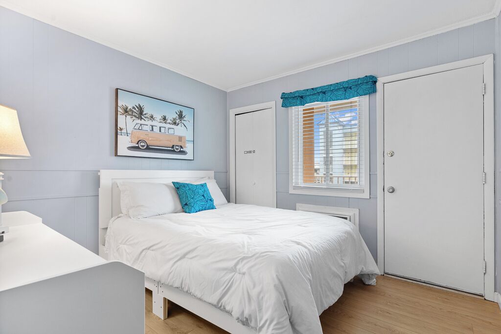 A bedroom with a bed, a bedside table, a window with blinds, a door, and a wall-mounted picture.