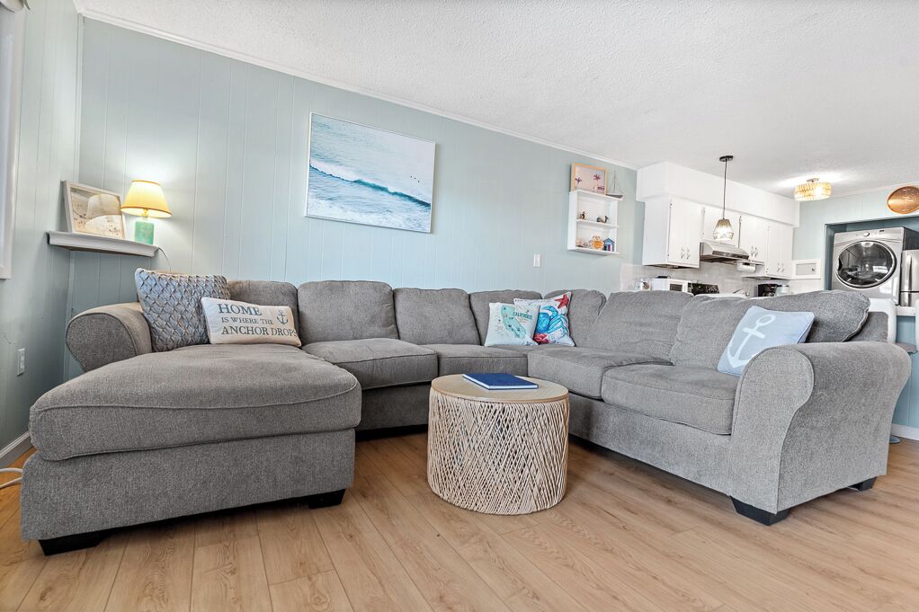 A living room with a grey sectional sofa, a round woven coffee table, and a kitchen in the background.