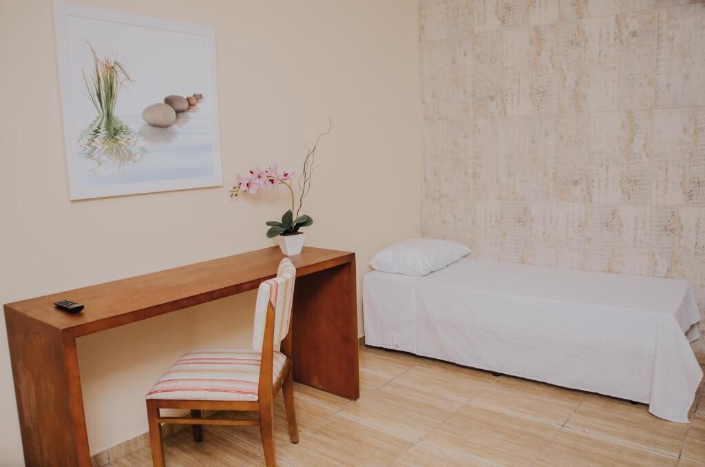 Une pièce comprenant un bureau en bois, une chaise, un lit recouvert d’une couverture blanche et une œuvre d’art fixée au mur représentant une plante et des rochers.