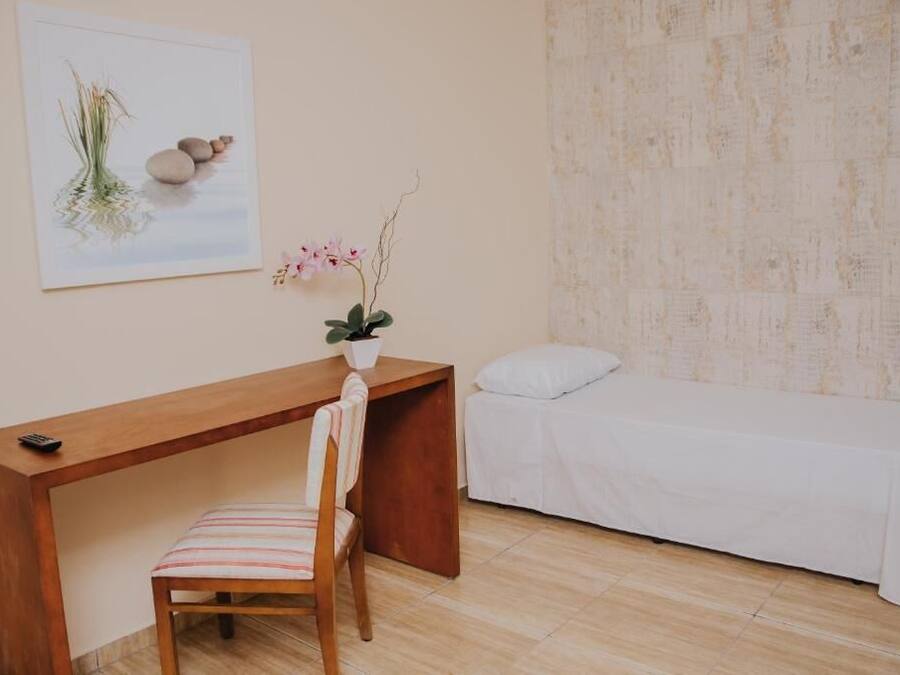 A room with a wooden desk, a chair, a bed with a white bedspread, and a wall-mounted artwork depicting a plant and rocks.