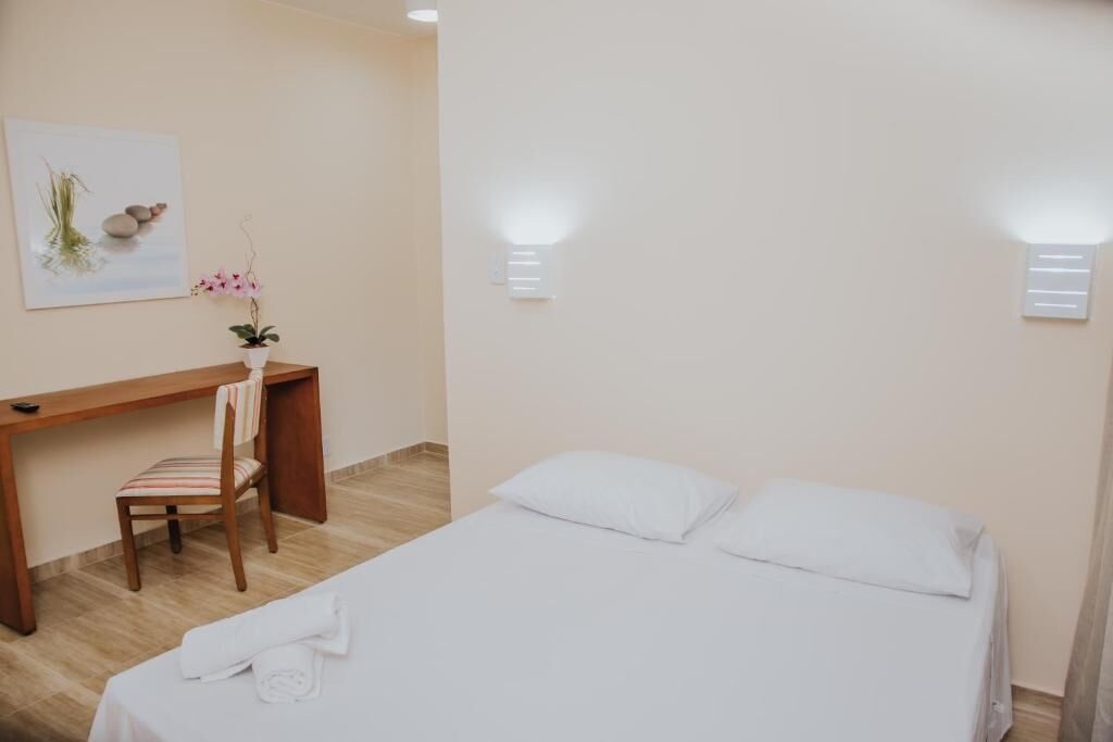 A hotel room with a bed, a desk, a chair, a potted plant, and a wall-mounted light fixture.