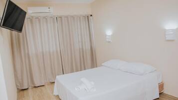 Een eenpersoonsbed met witte lakens, een flatscreen televisie aan de muur, een raam met beige gordijnen en een wandlamp.