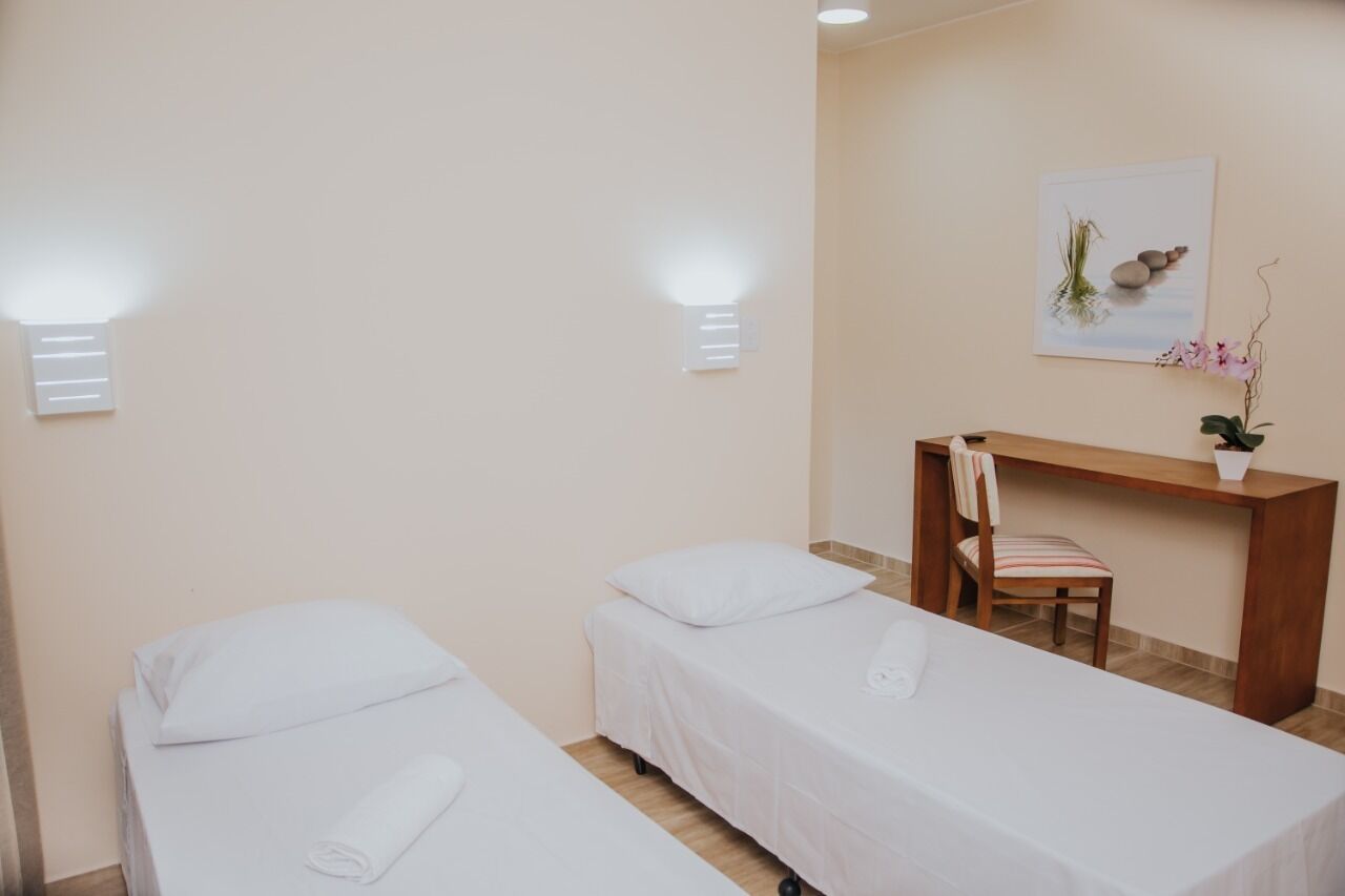 A room with two single beds, a wooden desk, a chair, and a wall-mounted light fixture.