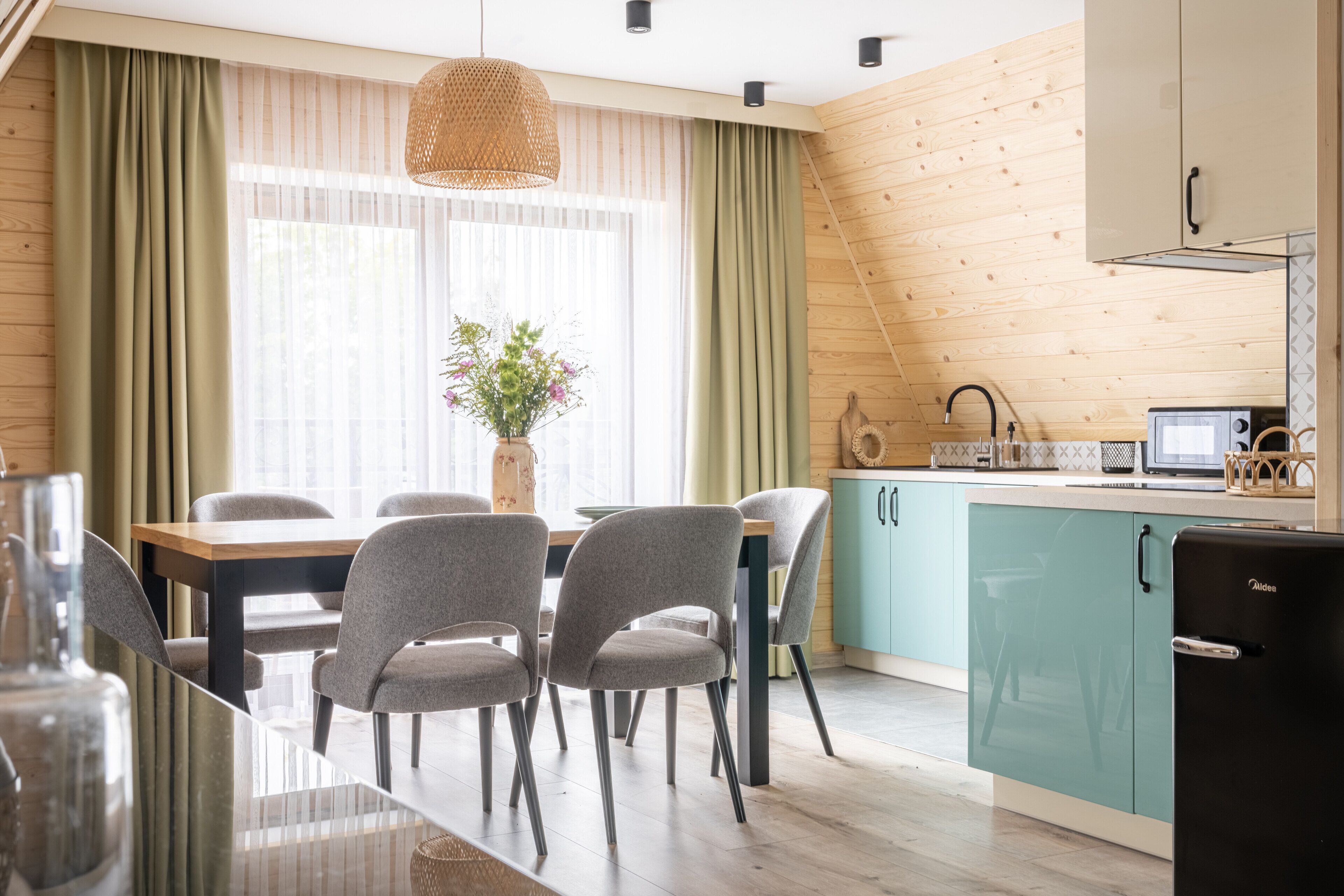 A modern kitchen with teal cabinets, a dining area with grey chairs, and a large window with sheer curtains.