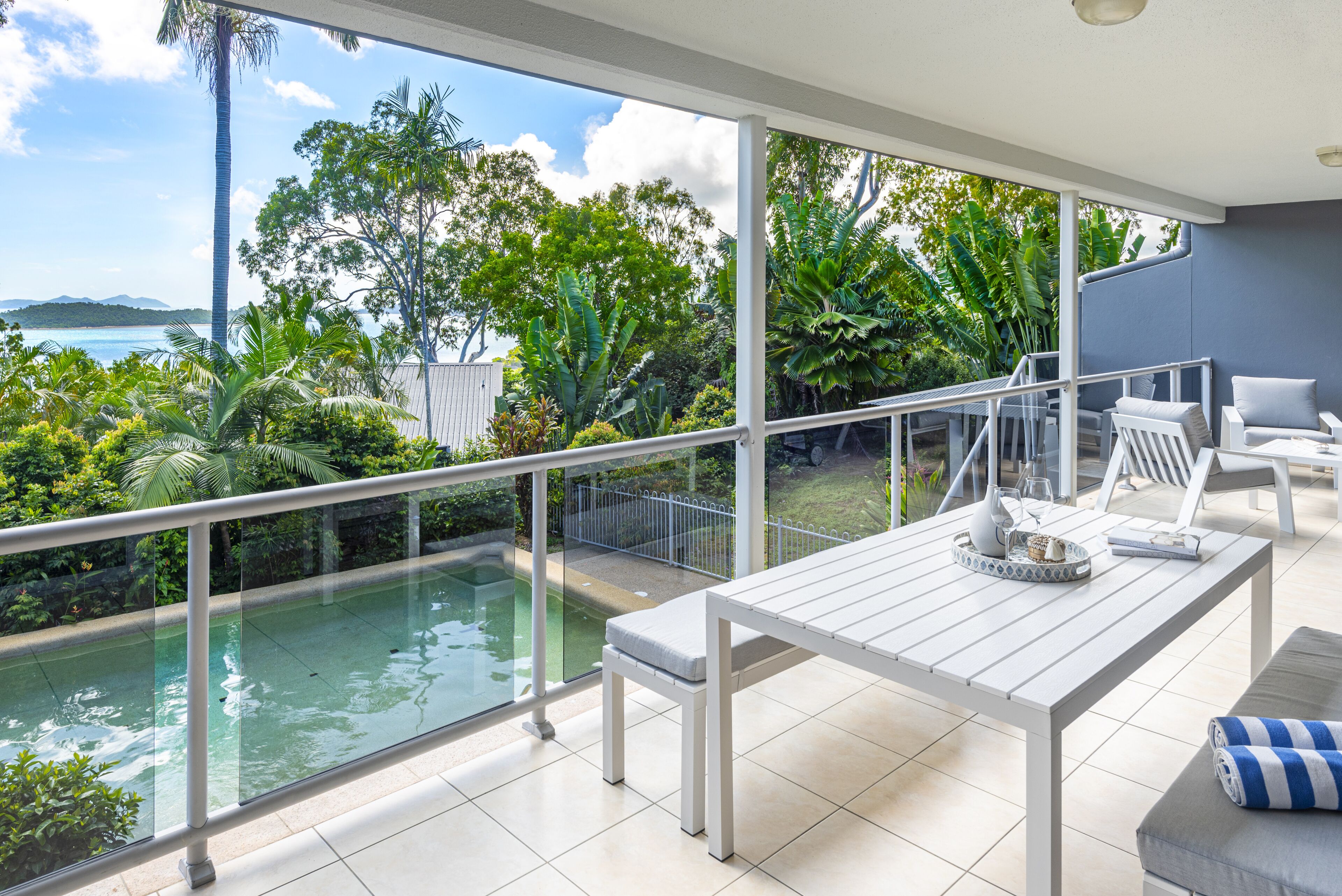 A balcony with a pool, white furniture, and a view of the sea and greenery.