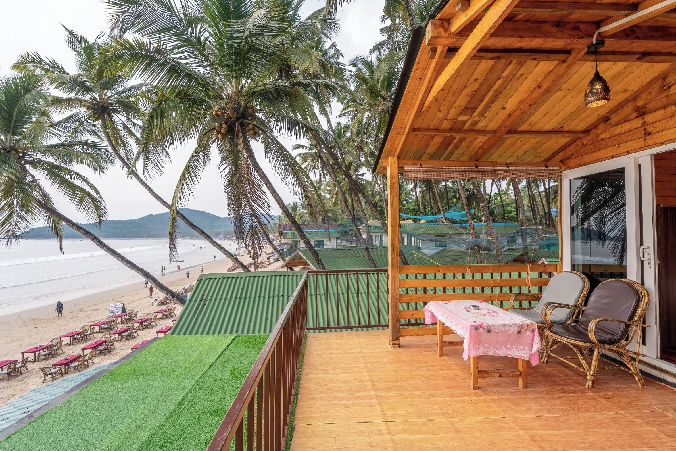 A wooden deck with a table and chairs, overlooking a beach with palm trees and a view of the sea.
