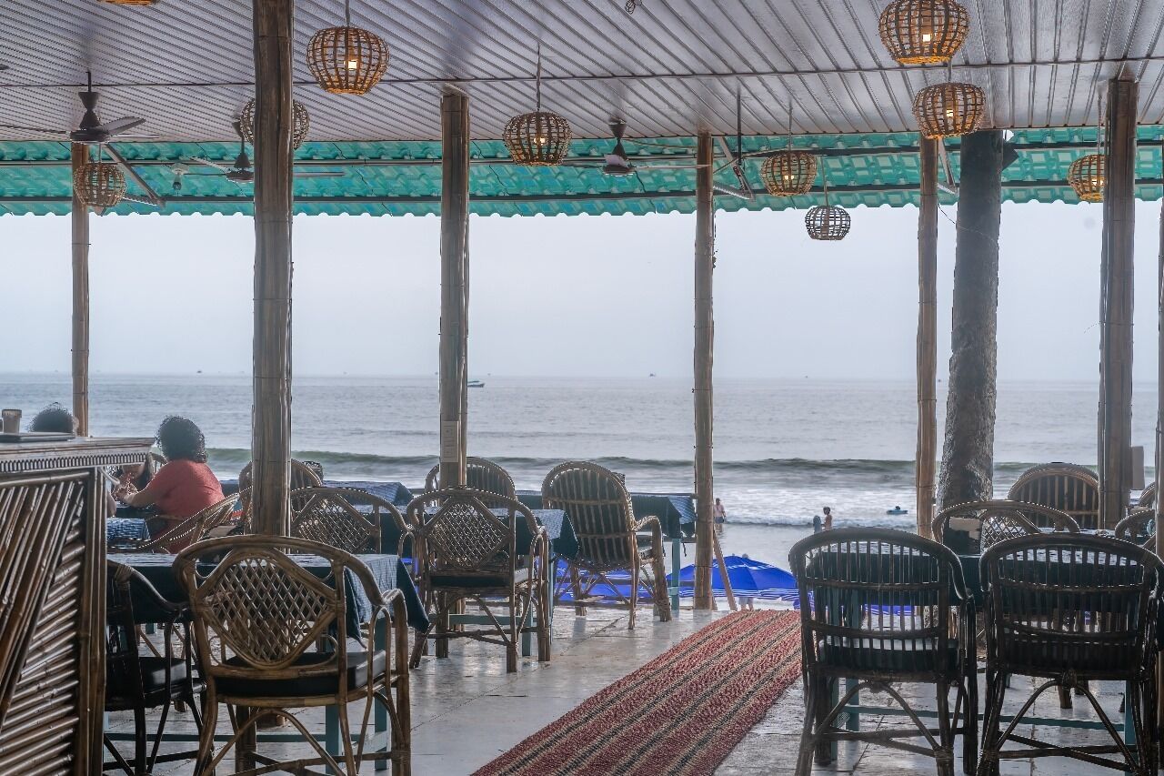 Un restaurante frente al mar con sillas y mesas de mimbre, una zona cubierta con techo verde y faroles colgantes.