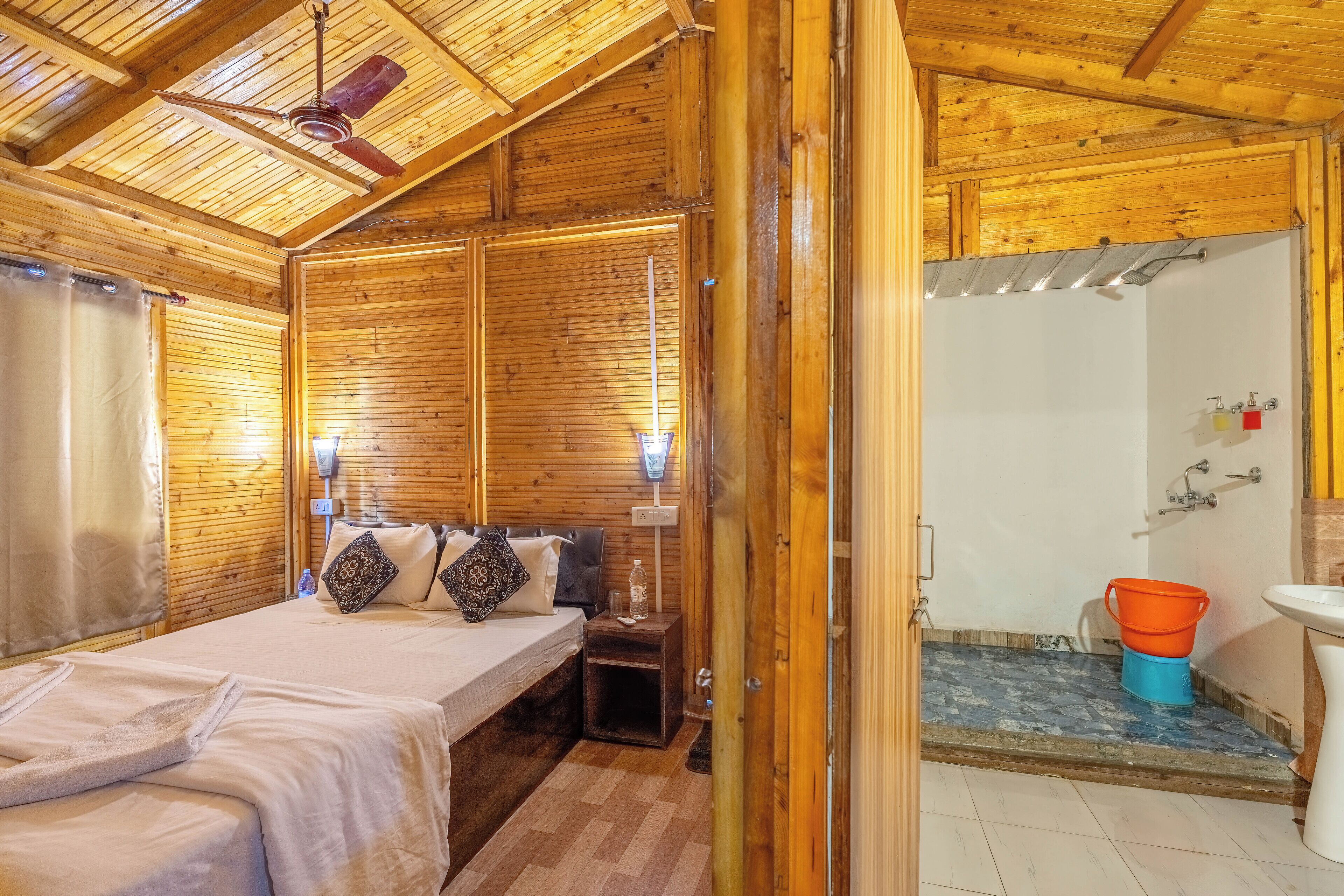 A wooden cabin with a bed, bedside table, and a bathroom with a sink and bucket.