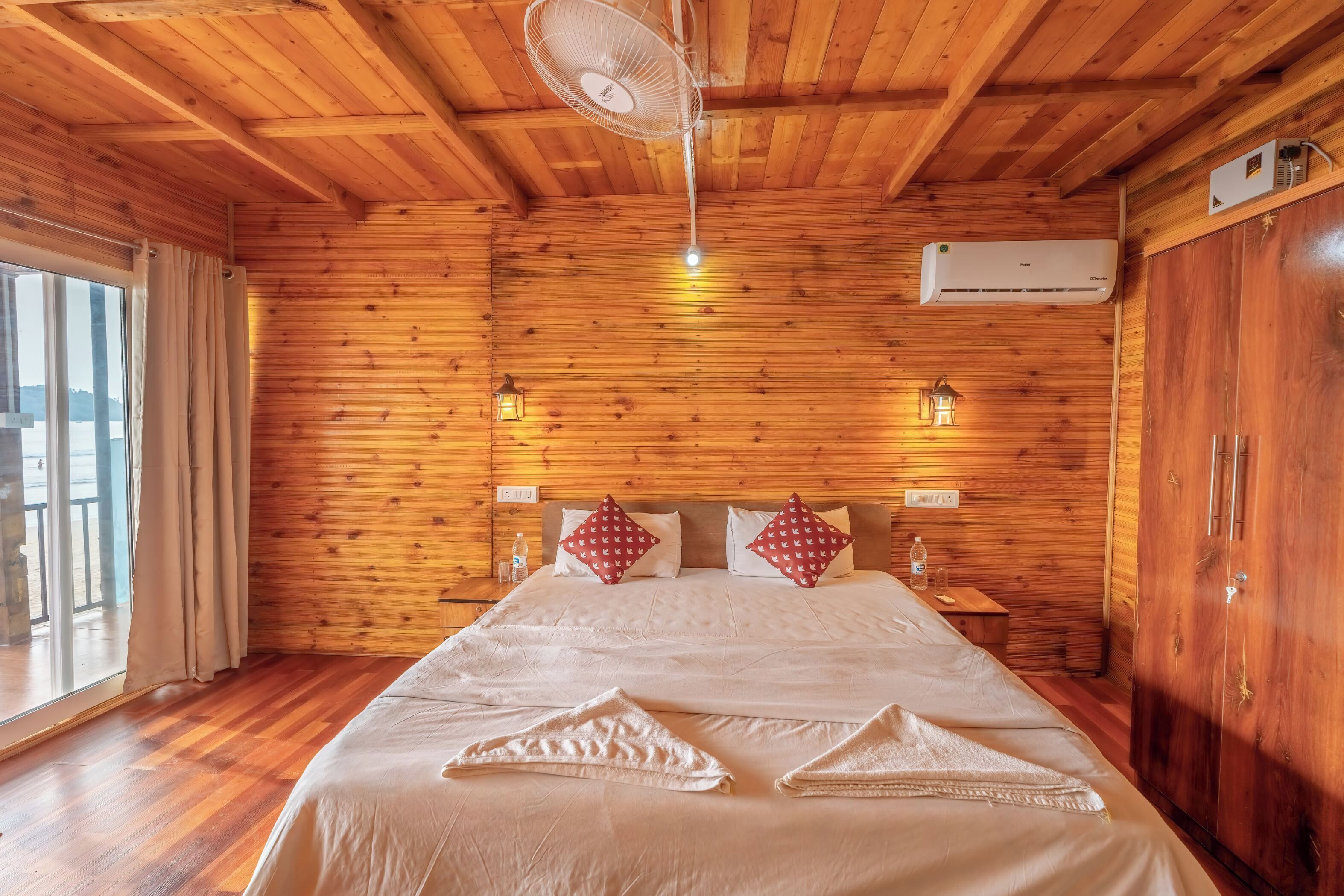 A wooden-paneled room with a bed, a fan, an air conditioner, and a window with curtains.
