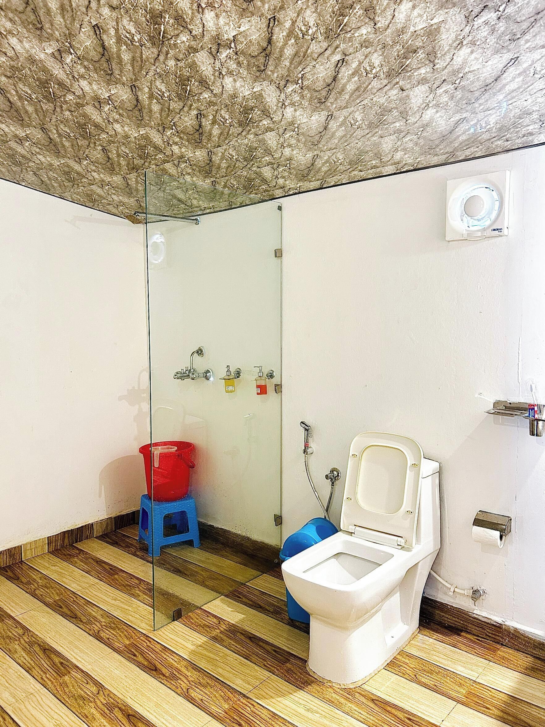 A bathroom with a glass partition, a white toilet, a red bucket, and a blue basin.