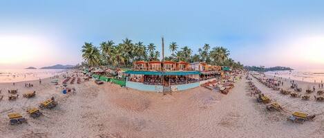 Un hotel frente a la playa con varias habitaciones, una piscina y una zona de playa con sombrillas y tumbonas.