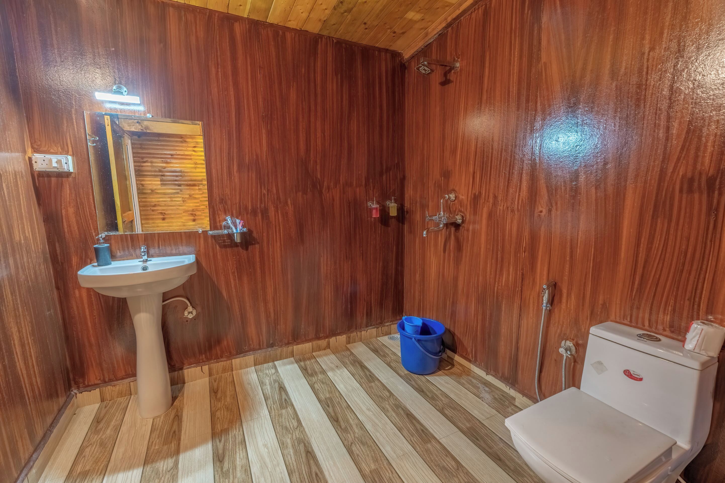 Une salle de bain avec des murs en bois, un lavabo, des toilettes et un seau bleu.