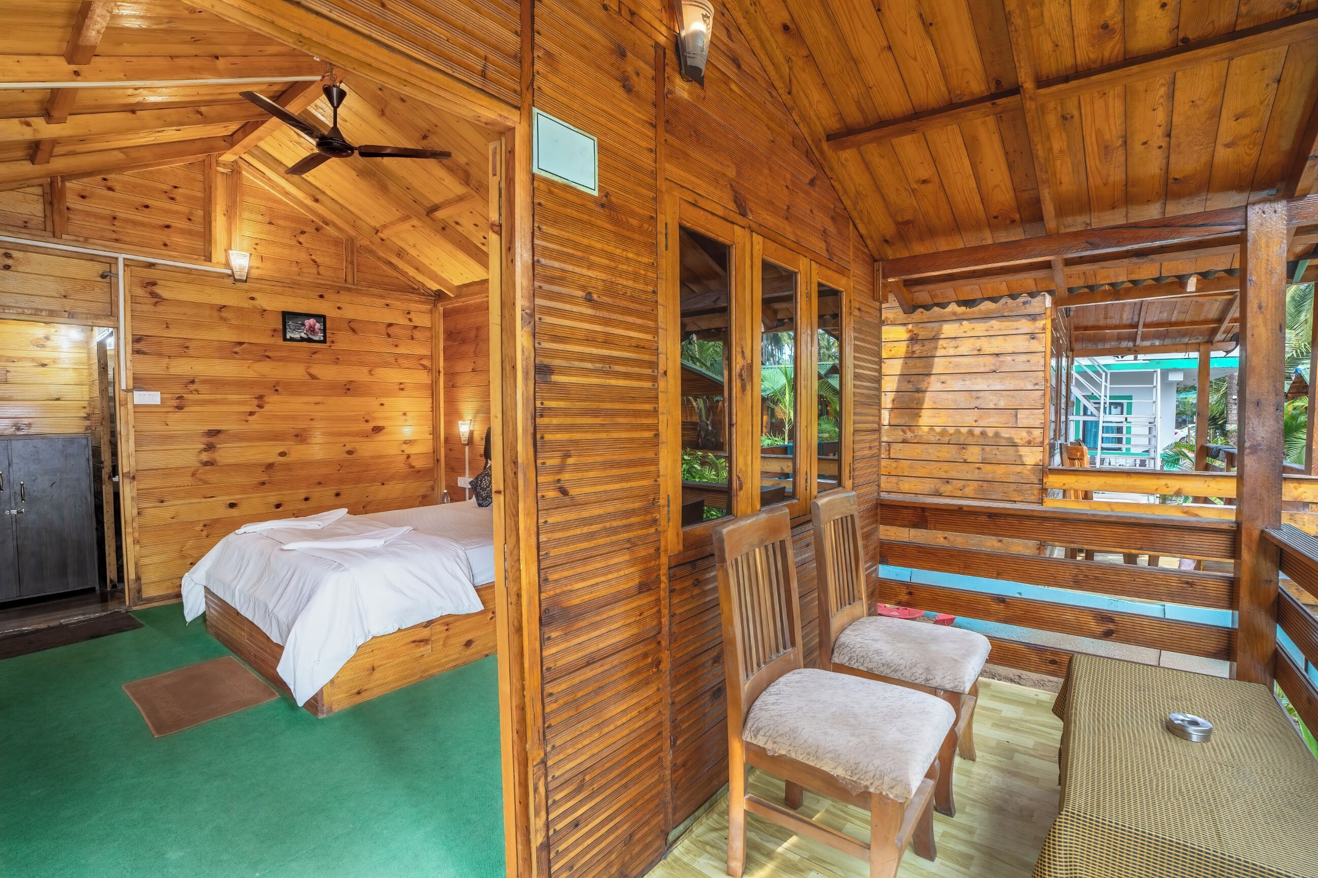 A wooden cabin with a bed, two chairs, a ceiling fan, and a view of the outside.