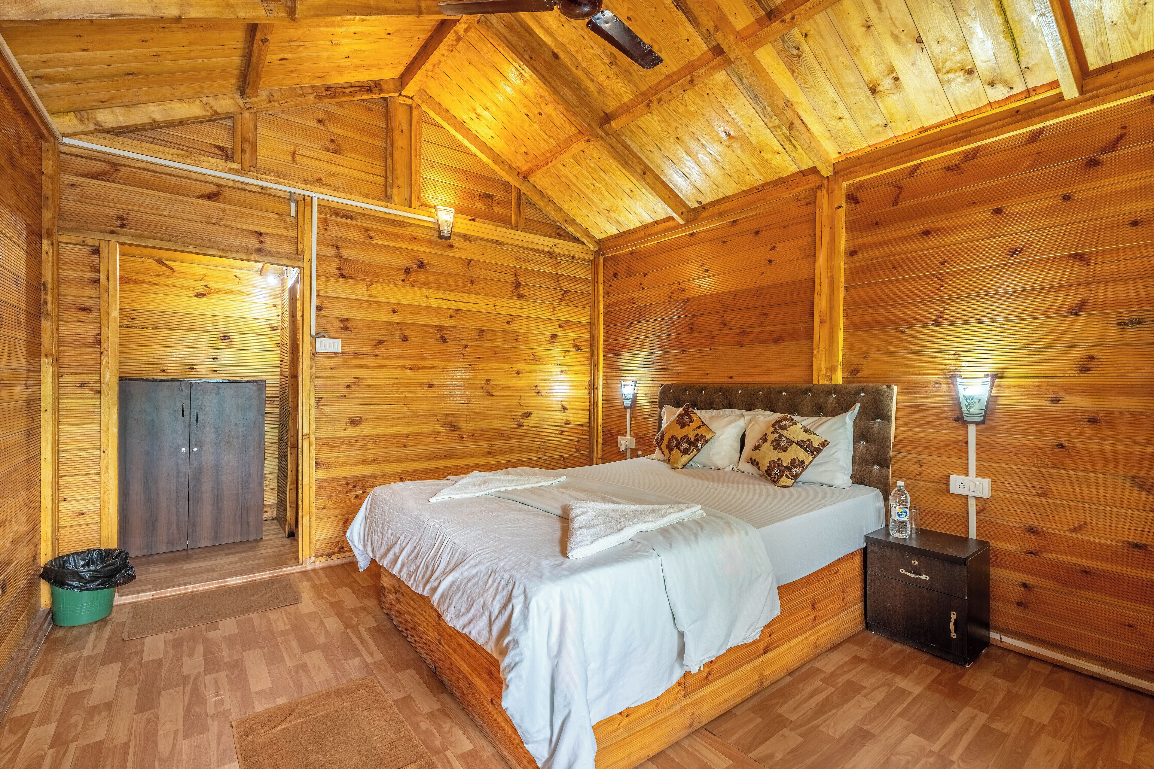 A wooden cabin room with a bed, nightstand, and a door leading to another room.