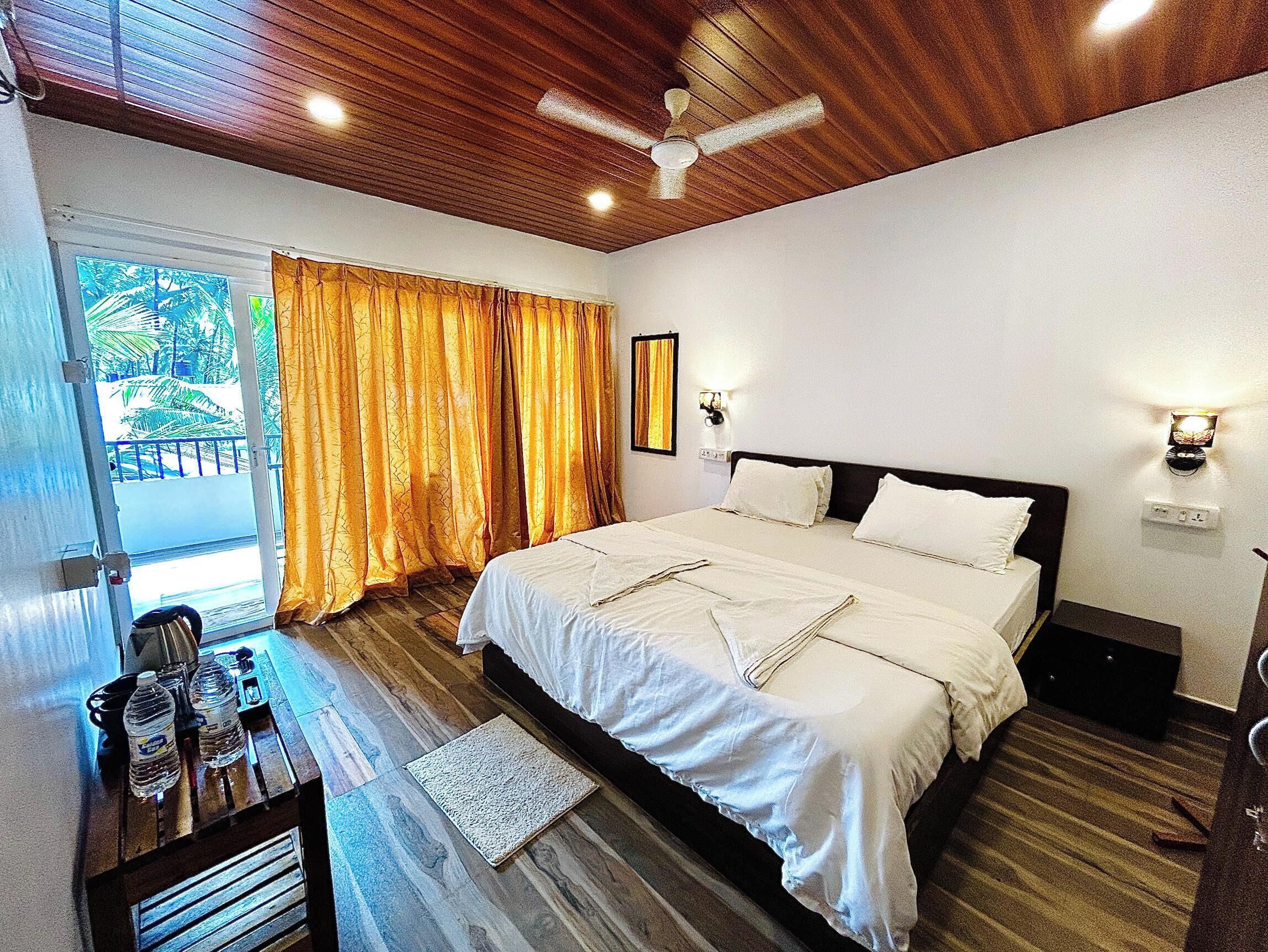 A hotel room with a bed, a desk with water bottles, a ceiling fan, and a view of the outdoors.