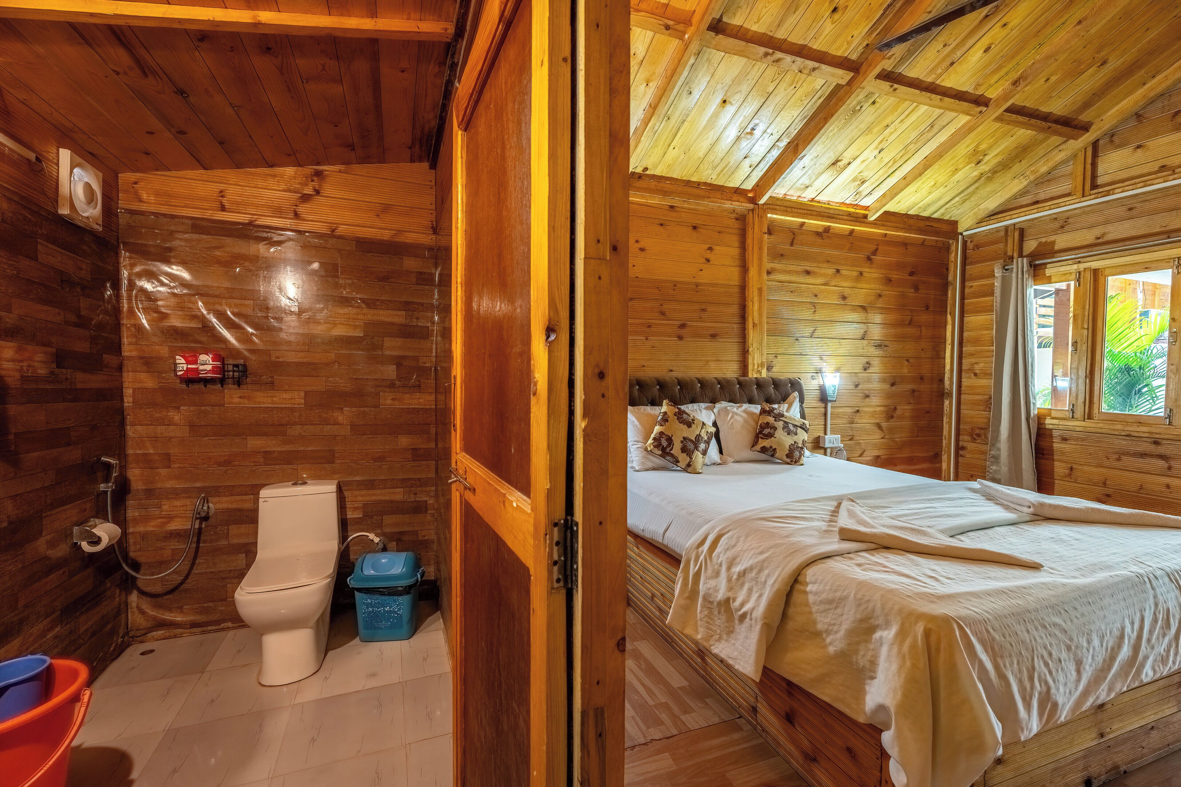 A wooden cabin with a bathroom and a bedroom. The bathroom has a toilet, a shower, and a bucket. The bedroom has a bed with pillows and a nightstand.