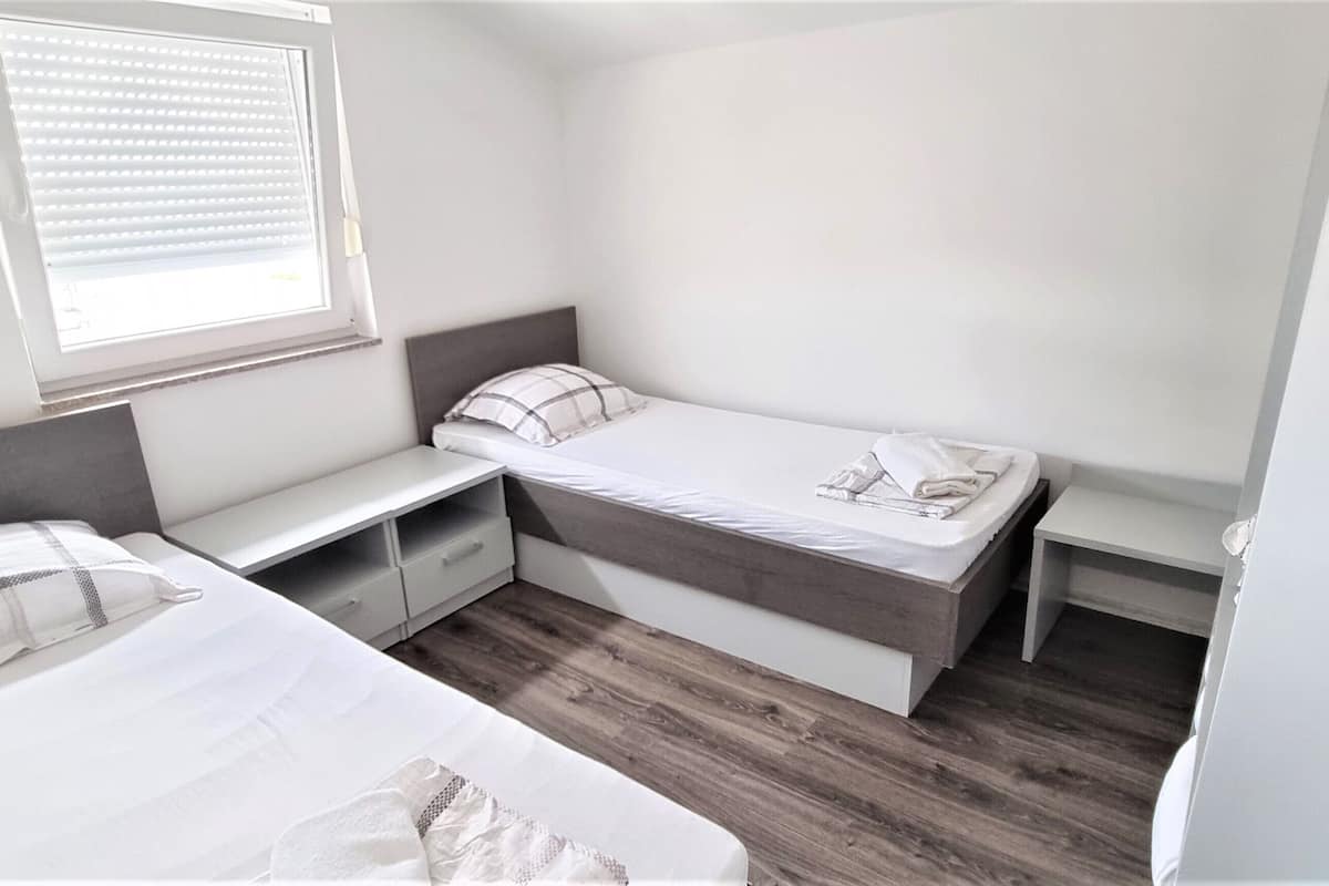 A small, well-lit bedroom with two single beds, a bedside table, and a window with blinds.