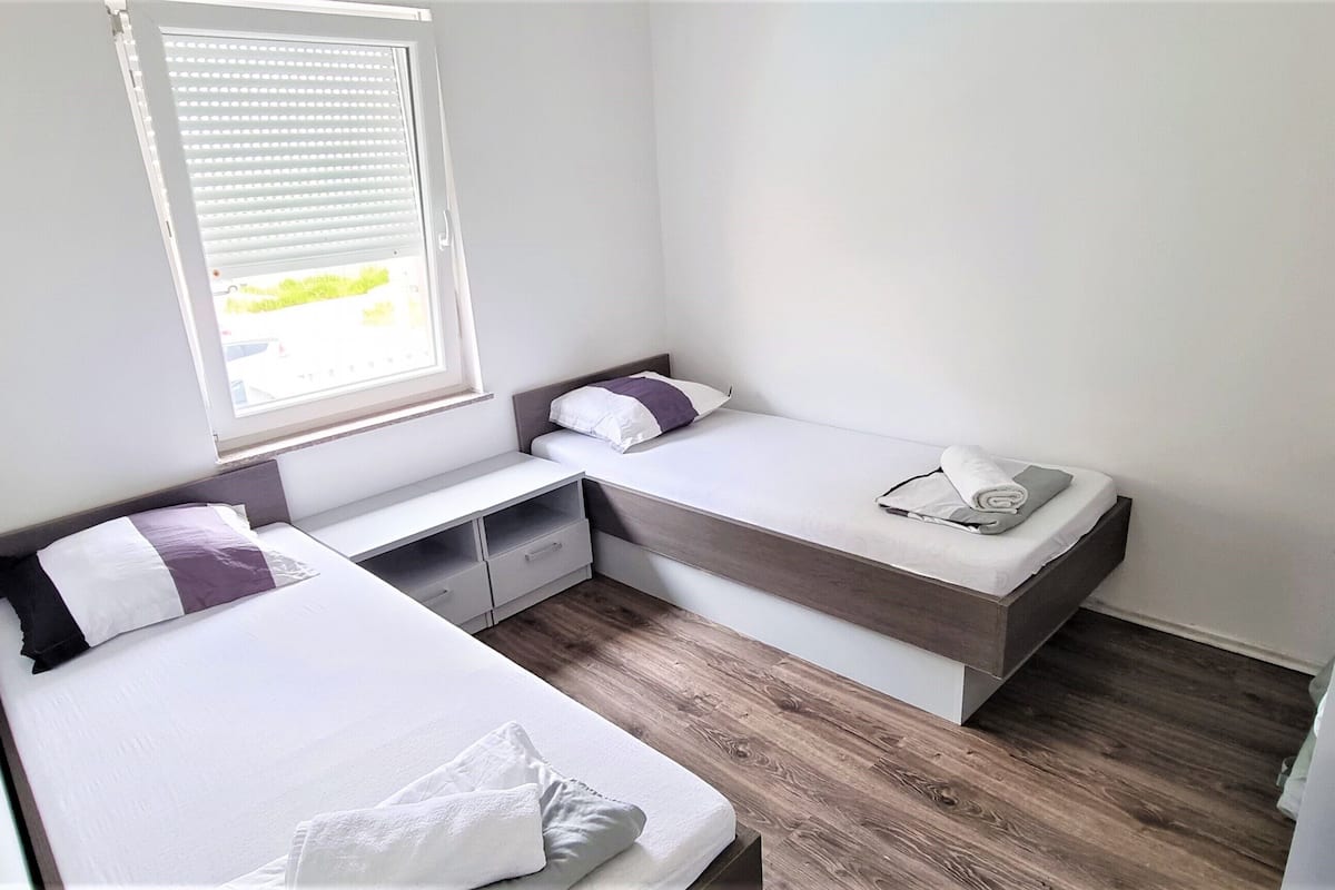 A compact bedroom with two beds, a small desk, and a window with blinds.