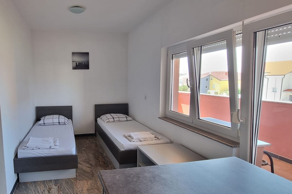 A dorm-style room with two single beds, a small table, and a window with a view of buildings.
