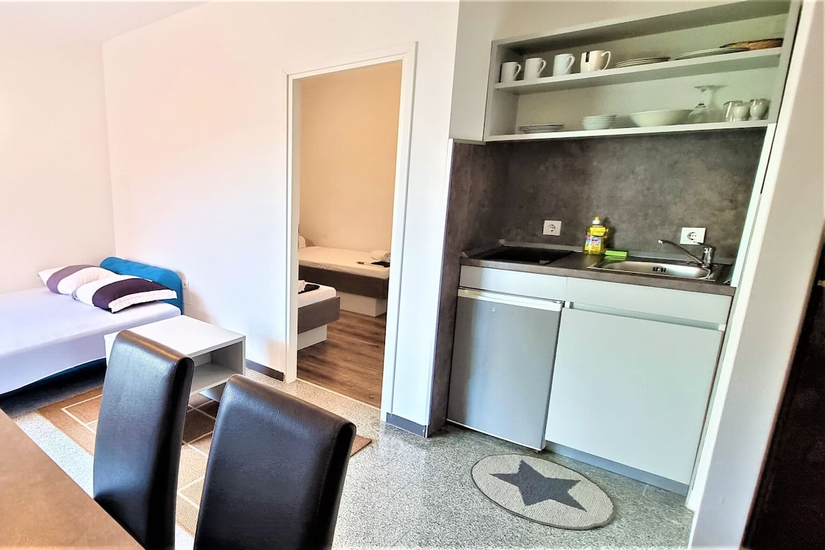 A compact room with a bed, a small kitchenette, a dining area, and a view of the cityscape.
