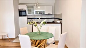 A modern kitchen with white cabinets, a built-in oven, and a microwave. There is a dining area with a round table and white chairs. - Tiffany Novalja (Novalja)