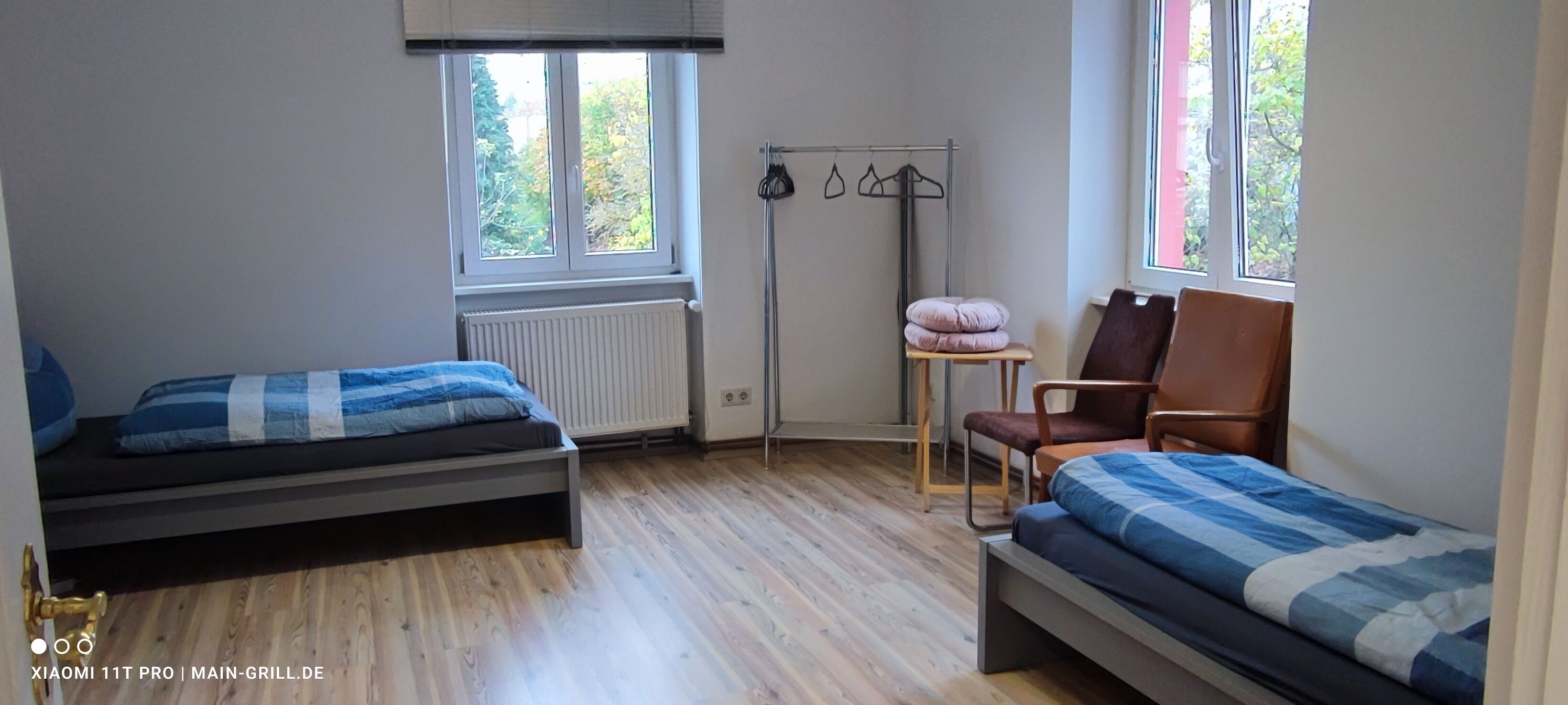 Un dormitorio con dos camas individuales, una silla, un radiador y una ventana con vistas a árboles.