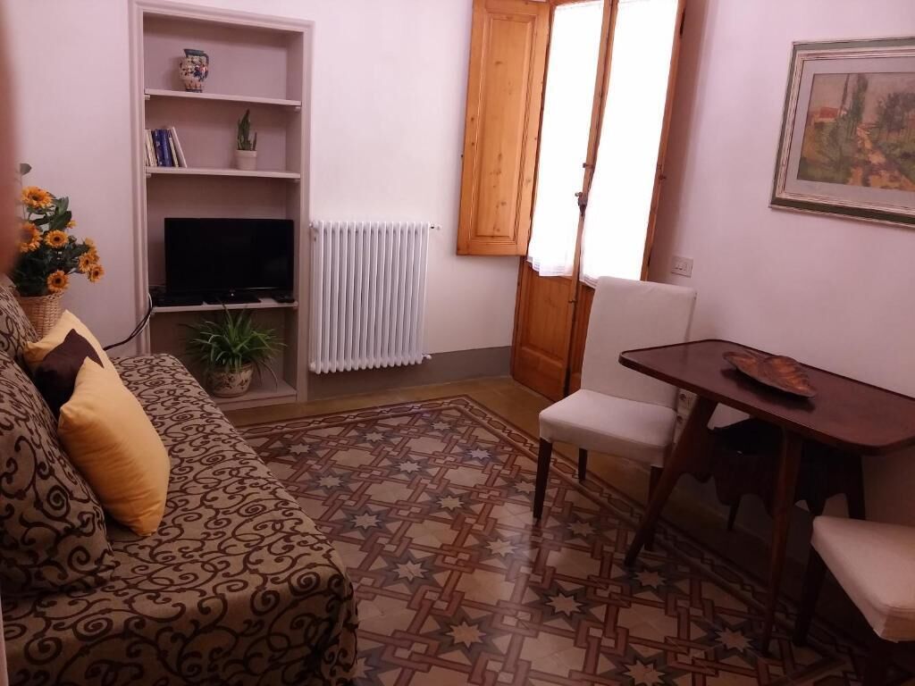 Una sala de estar con un sofá estampado, un televisor sobre una estantería, un radiador, una puerta de madera y un cuadro en la pared.