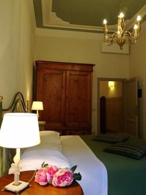 Quarto de hotel com cama, guarda-roupa de madeira, candelabro e mesa de cabeceira com abajur e flores.