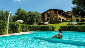 Una piscina con una persona sentada en el agua, rodeada de vegetación y un edificio de fachada marrón y ventanas arqueadas.