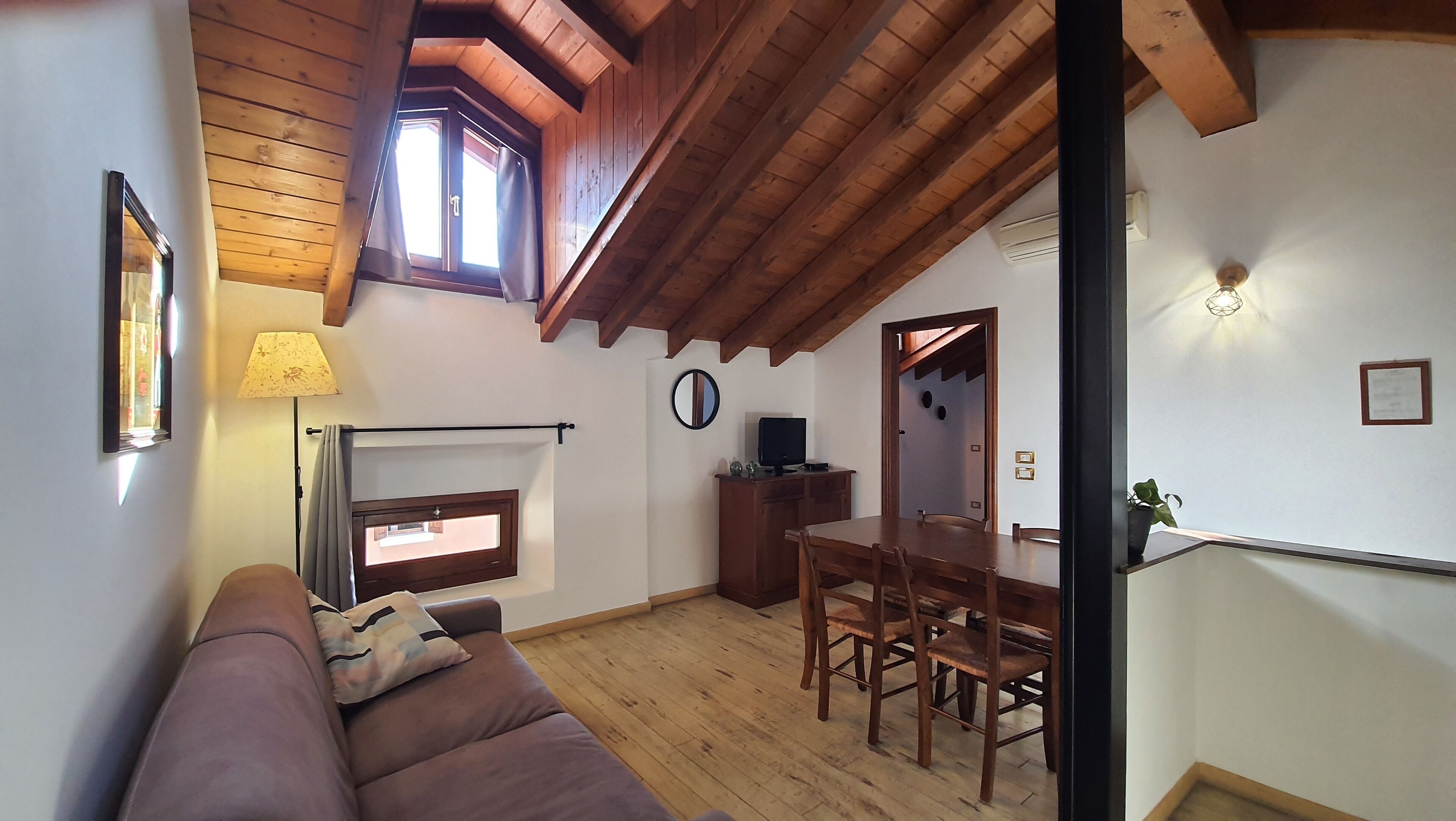 A cozy living room with a wooden ceiling, a sofa, a dining table with chairs, a TV, and a window with curtains.