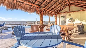 Um alpendre à beira-mar com telhado de palha, uma mesa redonda e cadeiras Adirondack azuis.